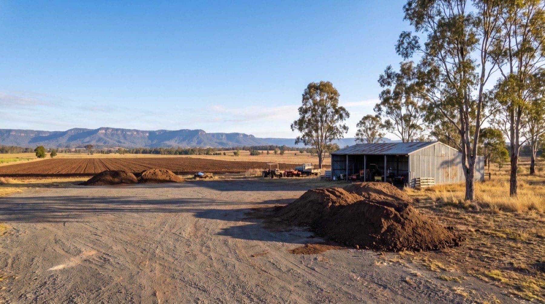 A working rural yard with a graded gravel hardstand, earth mounds and a machinery shed