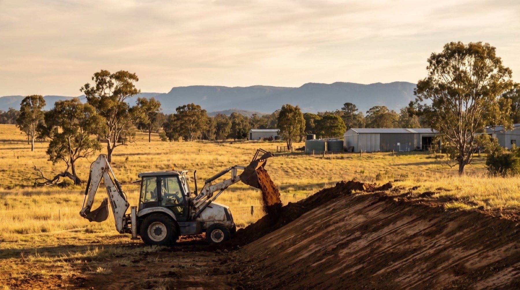 A backhoe cutting into a low rise on rural acreage in late afternoon light