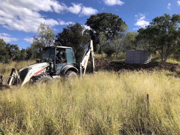 A backhoe working beside a corrugated water tank in long dry grass