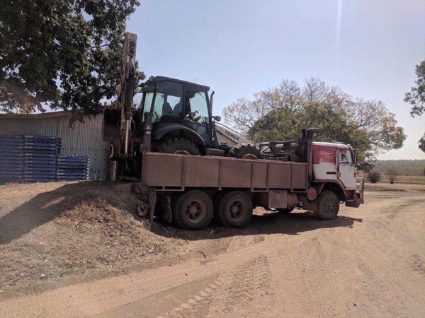 The backhoe loaded onto the tip truck beside a machinery shed