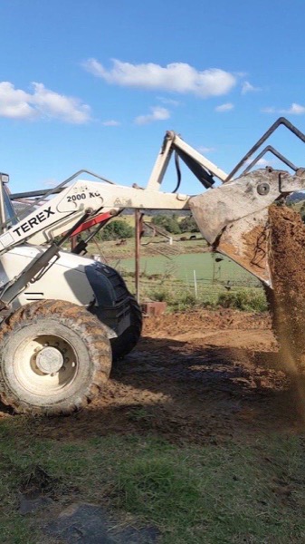 A loader bucket tipping a load of soil beside a paddock
