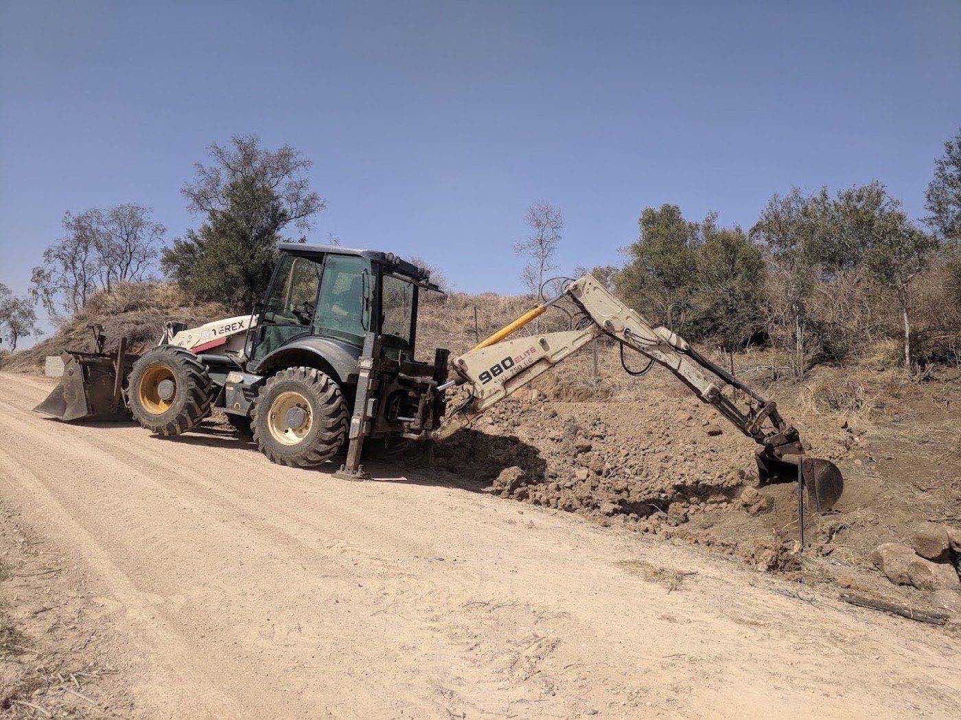 A backhoe cutting a trench into the shoulder of a gravel road on rural acreage