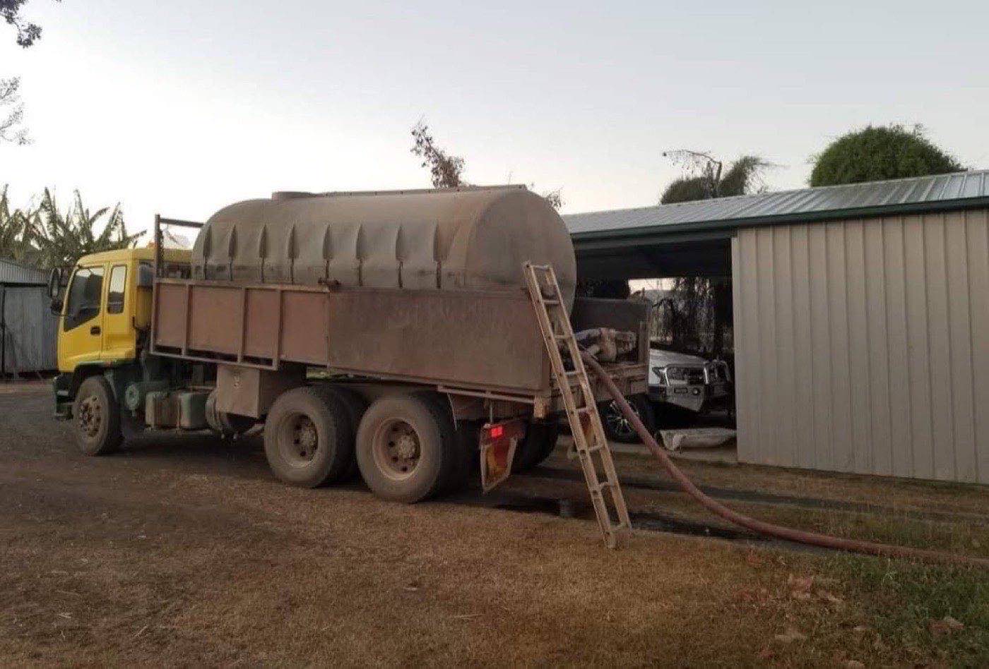 The water truck parked at a shed with the delivery hose running to a tank