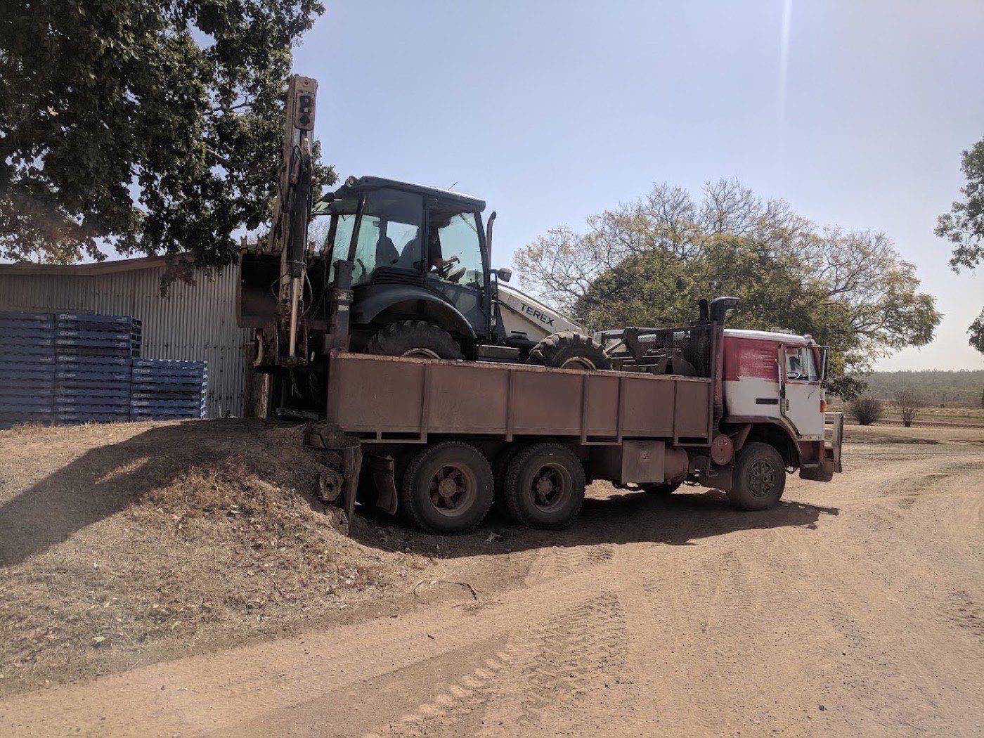 The backhoe loaded onto the tip truck beside a machinery shed