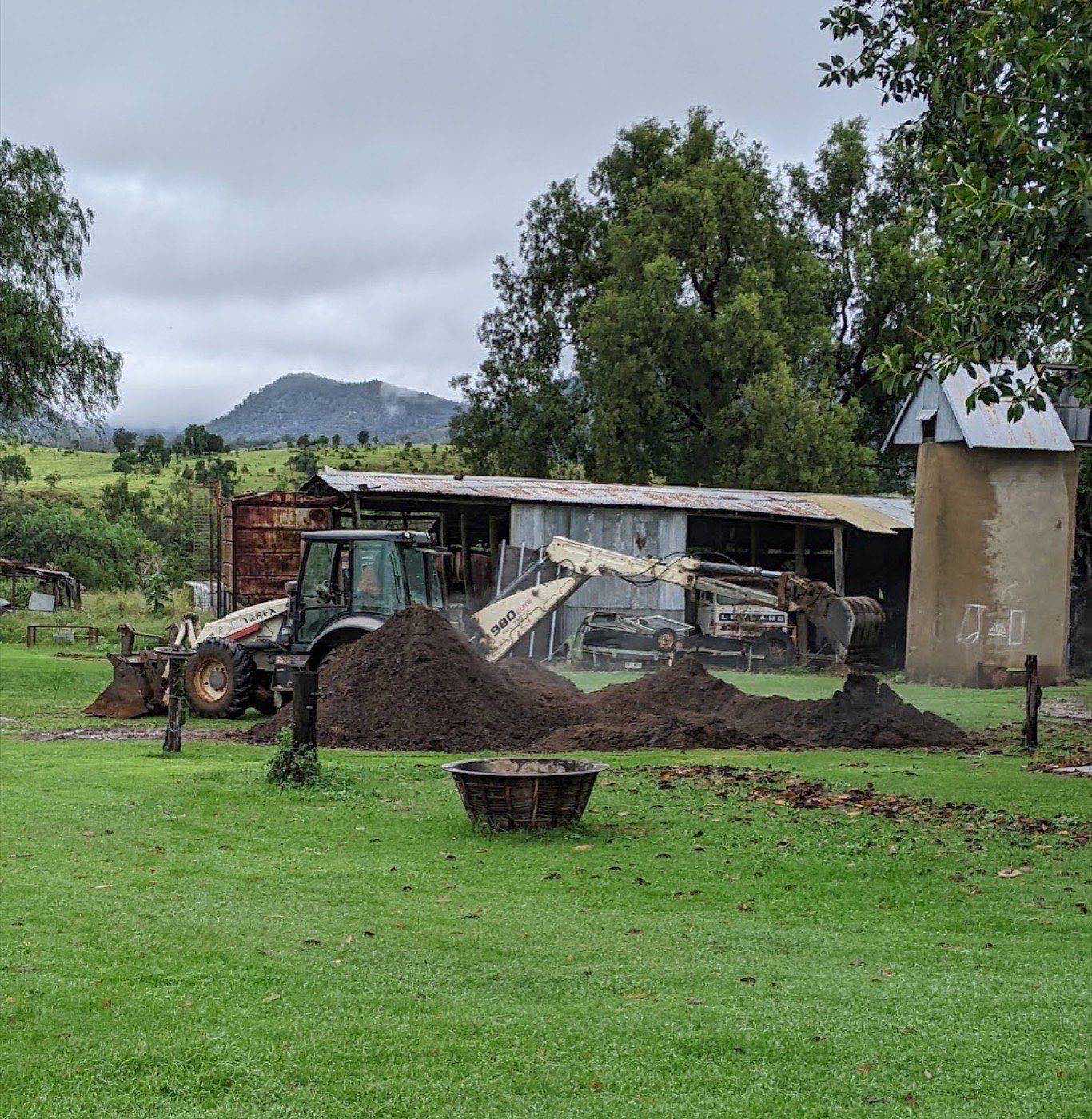 A backhoe beside heaps of freshly turned soil on a lawn in front of a farm shed