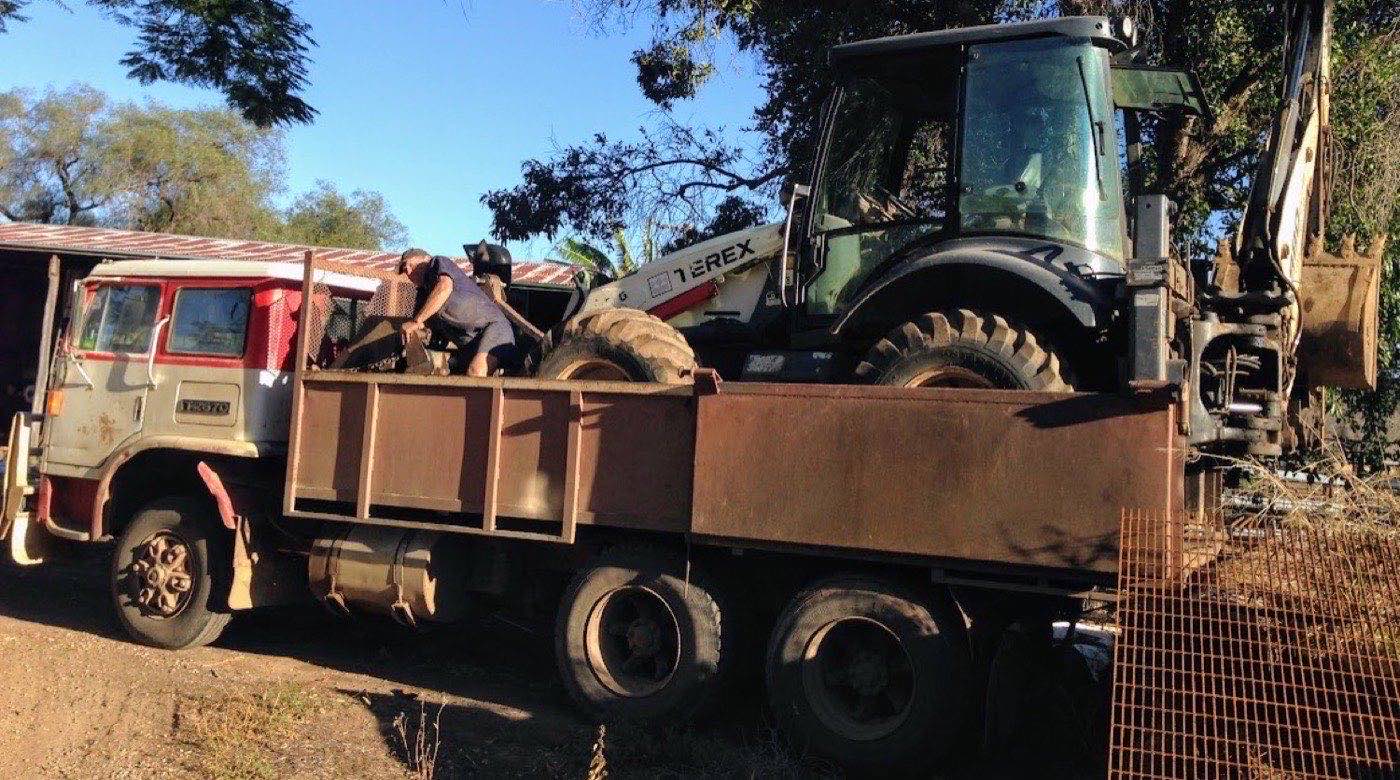 Gear being loaded onto the tip truck tray beside the backhoe in late afternoon light